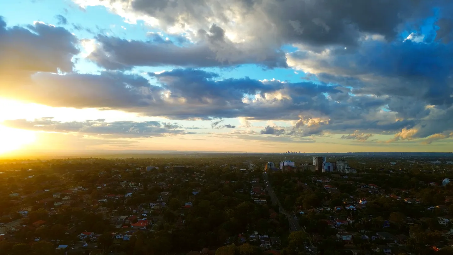 An aerial view over a wide city at golden hour, low sun breaking through cloud over neighborhoods and small commercial streets